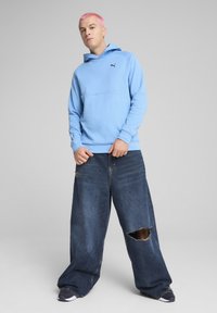 Sudadera azul claro con un logo negro, combinada con unos pantalones vaqueros oscuros de gran tamaño que presentan un desgastado rasgado. La sudadera tiene un bolsillo tipo canguro en la parte delantera.