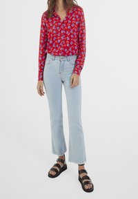 Chemise rouge à manches longues avec motif floral, associée à un jean évasé bleu clair et des sandales noires à brides avec des semelles épaisses.