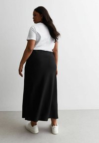 Jupe maxi noire en tissu texturé, portée avec un t-shirt blanc et des baskets blanches. Vue de dos, mettant en avant une silhouette lisse.