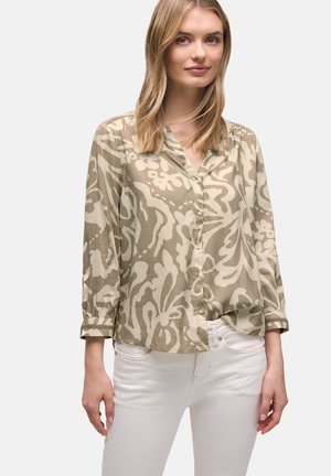 Femme blonde portant un chemisier à manches longues à boutons beige et crème à motifs, et un jean blanc, debout devant un fond uni.