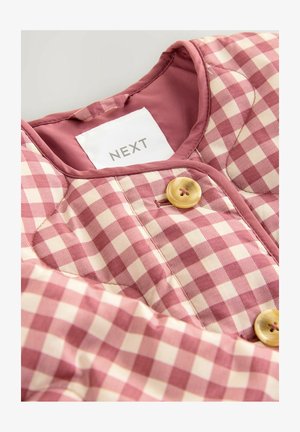 Vaate valmistettu vaaleanpunaisesta ja kuudelta kermalta ruudullisesta kankaasta, jossa on pyöreä pääntie, napitus ja valkoinen etiketti, jossa lukee "NEXT".