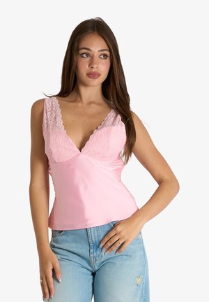 Donna con capelli lunghi castani che indossa un top senza maniche rosa con bordi in pizzo e jeans blu strappati, posando con una mano sui fianchi.
