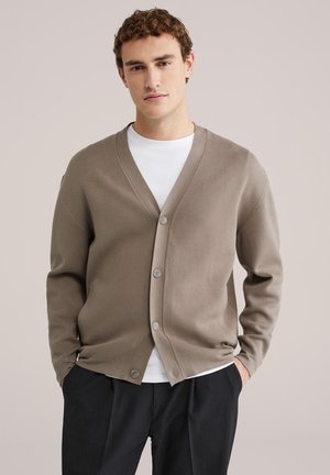 Bruine cardigan met een V-hals, knoopsluiting en lange mouwen. Gedragen over een wit shirt met ronde hals, met een gladde, zachte textuur.