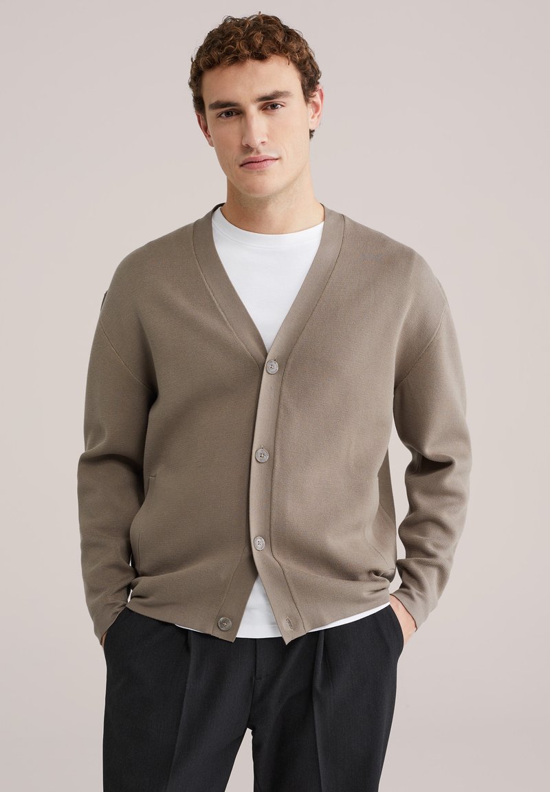 Pruun kardigan v-kaelusega, nööpidega ja pikkade varrukatega. Kanda valge ümarakaelusega särgi peal, millel on sile ja pehme tekstuur.