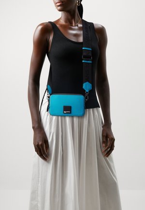 Vrouw die een zwarte mouwloze top en een witte rok draagt, met een blauwe crossbodytas met een brede zwarte riem en een metalen merkembleem.