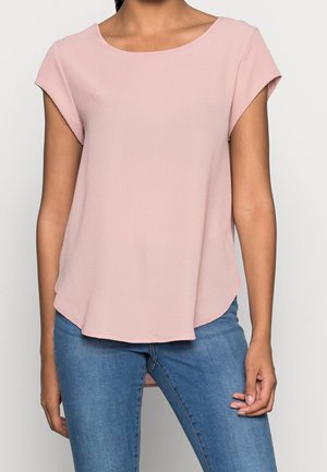 Femme portant un chemisier rose clair à manches courtes avec un col arrondi et un jean bleu, debout devant un fond neutre.