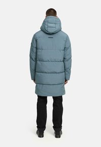 Light blue puffer coat with a hood, featuring a quilted design, elongated cut, and a subtle back logo detail. Worn with black pants and boots.