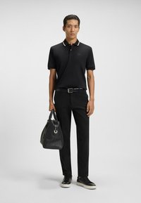 Czarna koszulka polo z kontrastowym kołnierzykiem, czarne spodnie, czarny pasek i czarne sneakersy. Trzyma strukturalną, czarną torbę na ramię.