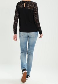 Schwarzes Oberteil mit langen Spitzenärmel und Schlüssel-Ausschnitt auf der Rückseite, kombiniert mit hellblauen Skinny-Jeans und schwarzen Sneakern. Glatte Stofftextur.