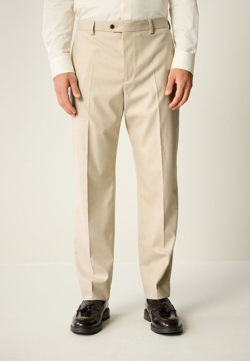 Homme portant un pantalon plissé beige, une chemise blanche et des mocassins à glands marron foncé, debout contre un fond uni.