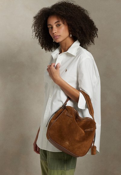 Woman with curly hair wearing a white shirt and green pants, holding a large brown suede shoulder bag.
