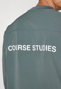 Grønn langermet skjorte med rund hals. Baksiden har teksten "KURS STUDIER" i store hvite bokstaver. Stoffet ser glatt ut.