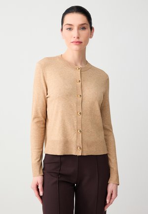 Kobieta ubrana w beżowy kardigan z długim rękawem z złotymi guzikami i ciemnobrązowe spodnie, stojąca na tle jednolitego jasnego tła.