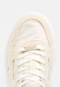 Gros plan de l’avant d’une basket beige en toile avec des lacets blancs et des coutures texturées autour de la zone des orteils.