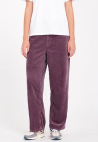 Pantalones de pana morados con un corte relajado, que presentan un diseño de rayas verticales y bolsillos laterales, combinados con una camiseta blanca sencilla y zapatillas.