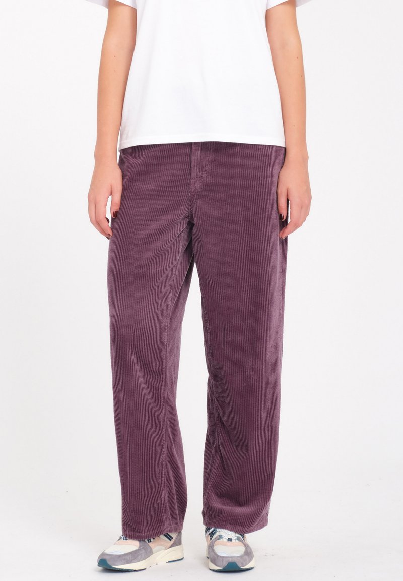 Pantalones de pana morados con un corte relajado, que presentan un diseño de rayas verticales y bolsillos laterales, combinados con una camiseta blanca sencilla y zapatillas.