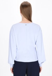Blusa azzurro chiaro con maniche lunghe e puff. Presenta una scollatura rotonda e una discreta chiusura sul retro con un piccolo accento metallico. Texture liscia.