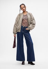 Casaco bomber acolchoado cinza-claro, blusa com padrão xadrez, jeans de perna larga azuis, botas castanhas de salto, segurando uma mala bordô.