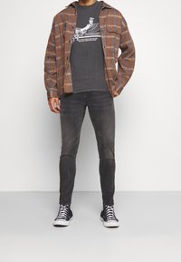 Man i brun rutigt jacka över svart grafisk t-shirt, mörkgrå jeans och svarta högstövlar som står mot en vit bakgrund.