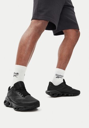 Nogi w czarnych sportowych butach z grubymi podeszwami i czarnych spodenkach, w połączeniu z białymi skarpetkami Reebok za kostkę, ustawione tak, jakby ktoś chodził lub pozował.