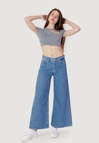 Top corto grigio a coste con maniche corte, abbinato a jeans in denim blu chiaro a gamba larga e sneaker bianche. Modella in posa con le braccia sollevate.