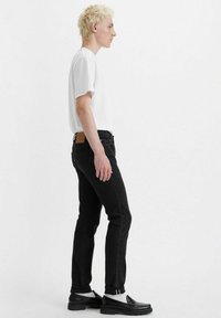 Jeans de mezclilla negra y una camiseta blanca, con un corte recto y un parche de cuero en la cintura trasera. El modelo lleva mocasines negros.