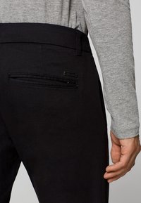 Pantaloni neri con una texture liscia, tasca posteriore con zip e un piccolo tag con il logo. Accompagnati da una maglietta grigia a maniche lunghe.