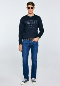 Jaunas vyras su akiniais nuo saulės, tamsiai mėlynu džemperiu su užrašu „BIG STAR“, mėlynomis džinsomis, diržu, laikrodžiu ir mėlynomis sportbačiais, stovintis su viena ranka kišenėje.