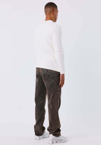 Hombre de espaldas vistiendo una camisa blanca de manga larga, jeans oscuros con remaches laterales y zapatillas deportivas blancas sobre un fondo liso.