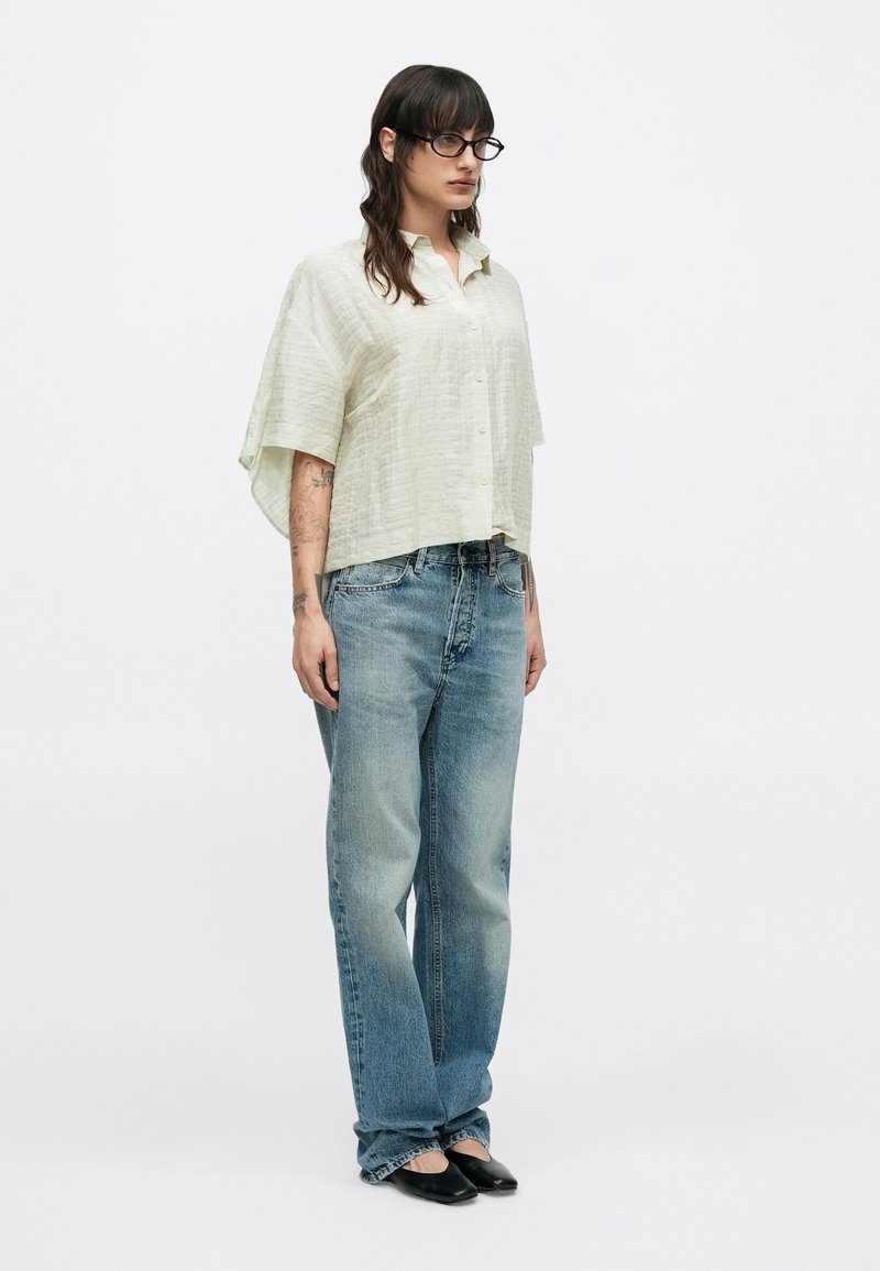 Lys grøn skjorte med knapper og en tekstureret finish, kombineret med løstsiddende blå denimjeans og sorte flade sko. Enkel, afslappet pasform med korte ærmer.