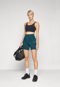 Svart sports-bh og høy midje shorts i teal med et subtilt mønster. Modellen har på seg svarte joggesko og holder en svart treningsbag.