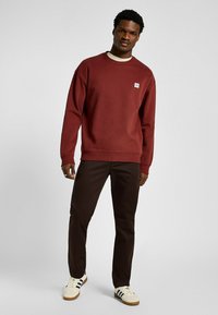 Bordowa bluza z okrągłym dekoltem o prążkowanym wykończeniu, z małą naszywką z logo. W zestawie z brązowymi spodniami i białymi trampkami z czarnymi paskami.