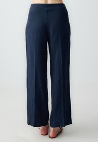 Navy linnen wijde broek met een soepele textuur en een hoge taille, met zijzakken en een vloeiende silhouet. Bruine sandalen op blote voeten.