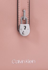 Borsa Calvin Klein in morbido materiale rosa chiaro con texture. Presenta una chiusura a lucchetto argentato e dettagli del logo ricamati in bianco.