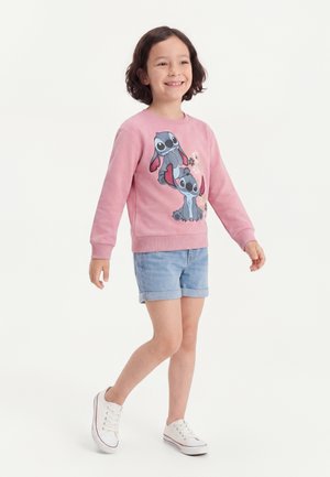 Enfant souriant portant un sweat-shirt rose avec des personnages de dessins animés, un short en jean et des baskets blanches, debout sur une jambe sur un fond uni.