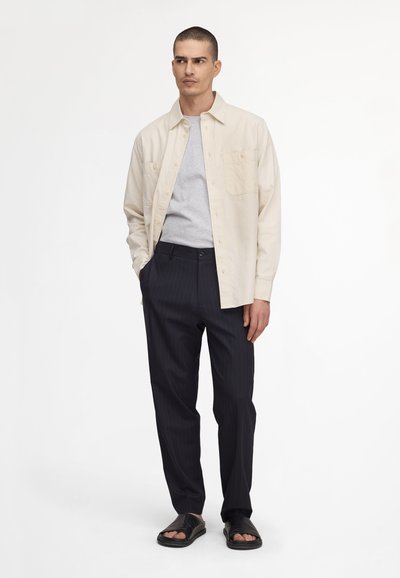 Homme portant une chemise beige boutonnée sur un t-shirt gris clair, pantalon noir à fines rayures et sandales noires ouvertes à l'avant, debout avec une main dans la poche.