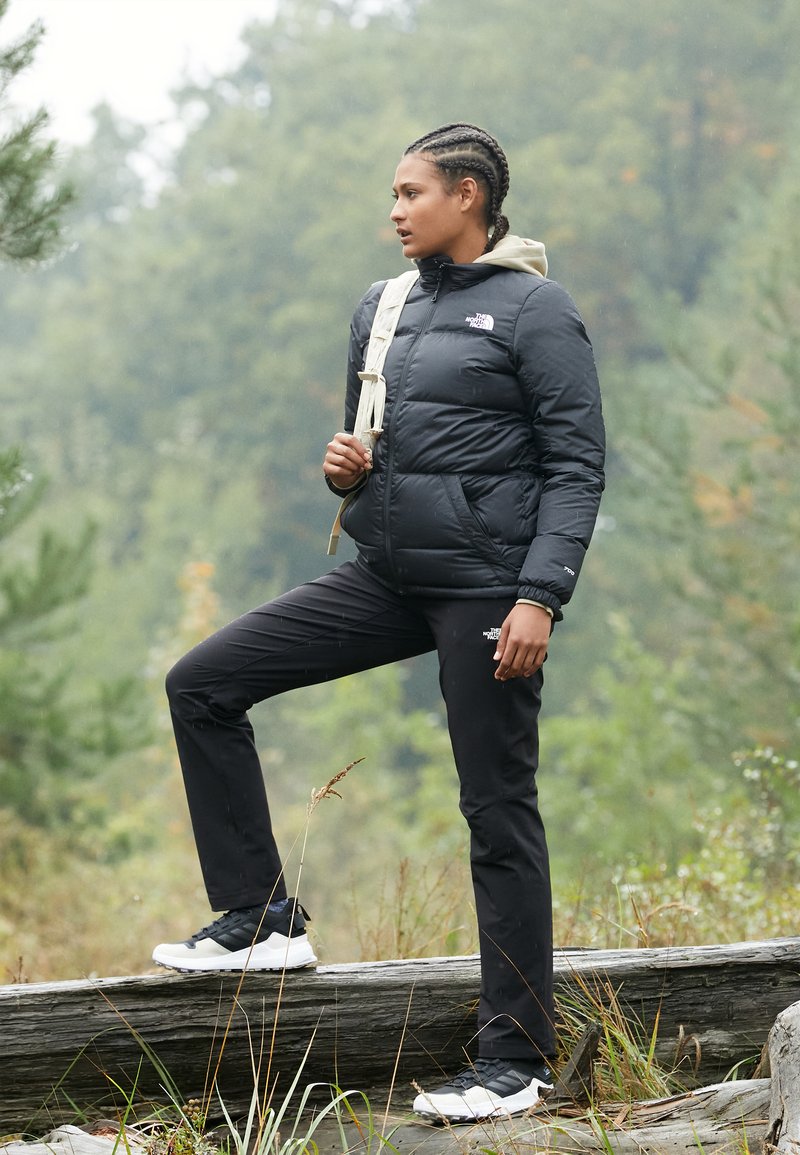Black puffer jacket with a hood, black pants, and white sneakers. Outdoor setting with greenery and logs in the background.