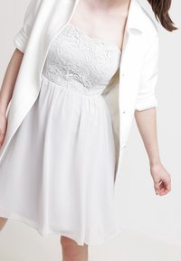 Robe blanche avec corsage en dentelle et jupe fluide, associée à une chemise blanche boutonnée. La robe présente un design sans bretelles et une texture légère.