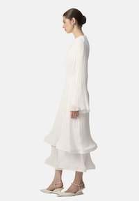 Robe plissée blanche longue avec des manches longues et un ourlet superposé. Associée à des chaussures à talons pointus de couleur claire. Texture lisse et fluide.