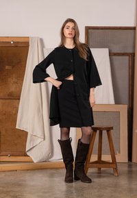 Czarny płaszcz zapinany na guziki z rozszerzanymi rękawami, zestawiony z dopasowaną spódnicą. Modelka nosi brązowe zamszowe kozaki za kolano. Miękka, teksturowana tkanina.