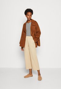 Manteau marron à double boutonnage porté sur un haut à rayures noir et blanc, associé à un pantalon large beige et des chaussures à enfiler en peluche couleur tan.