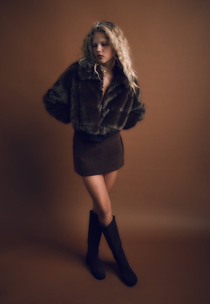Brun jacka i konstpäls, kombinerad med en brun mockakjol i kort modell och svarta knähöga stövlar, mot en varm brun bakgrund.