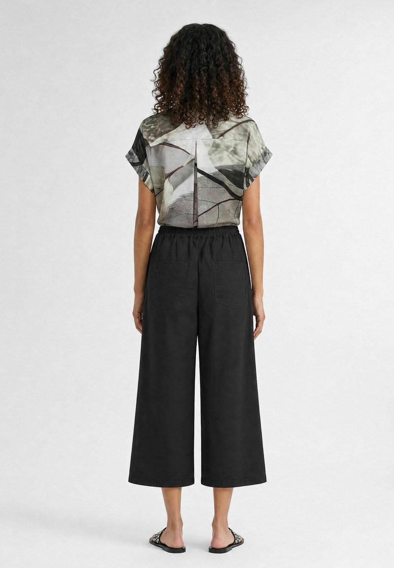 Femme aux cheveux bouclés porte un chemisier à manches courtes à motif abstrait, rentré dans un pantalon noir taille haute, large et court, et des sandales plates.