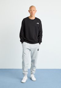 Mand står med hænderne i lommerne iført en sort sweatshirt, lysegrå sweatpants og hvide sneakers på et blåt gulv.