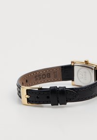Pulseira de relógio em couro preto com acabamento texturizado, apresentando uma fivela de metal na tonalidade dourada e um mostrador retangular prateado com gravação da marca.