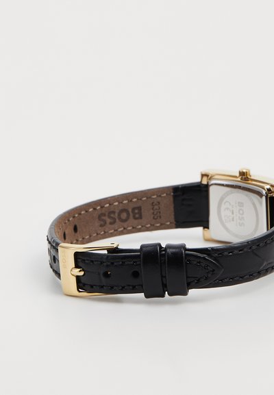 Bracelet de montre en cuir noir avec une finition texturée, dotée d'une boucle en métal doré et d'un cadran de montre rectangulaire en argent portant une gravure de la marque.