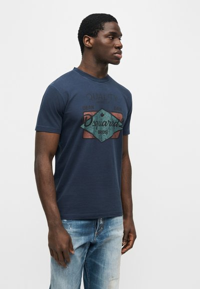 Νεαρός άνδρας που φοράει σκούρο μπλε t-shirt με σχέδιο και ανοιχτόχρωμο μπλε τζιν, στέκεται μπροστά σε απλό ανοιχτόχρωμο φόντο, κοιτάζοντας δεξιά.