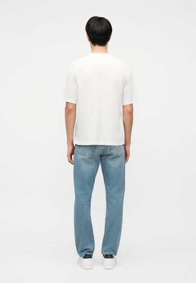 Homme debout, vu de dos, portant un t-shirt blanc uni à manches courtes, un jean bleu clair et des baskets blanches sur un fond blanc.