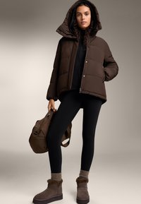 Bruine pufferjack met een capuchon, zwarte leggings en bruine laarzen met bontvoering. Houdt een bruine handtas vast, tegen een neutrale achtergrond.