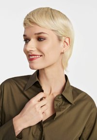 Oliwkowa, zapinana na guziki koszula z kołnierzykiem. Model lekko trzymający kołnierzyk, ukazując subtelną fakturę. Wielokrotne kolczyki w uszach.